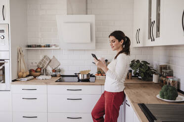 Smiling woman using smart phone in kitchen at home - EBBF08151