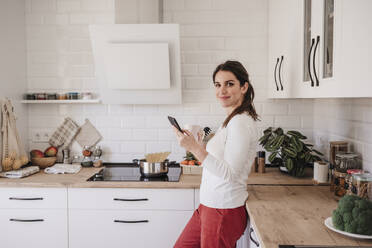 Smiling woman with mobile phone standing by counter in kitchen - EBBF08152