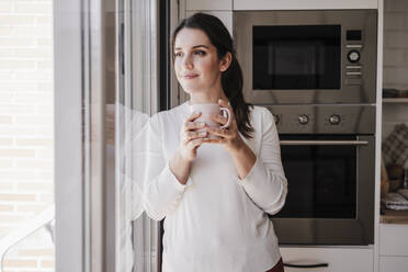 Nachdenkliche Frau mit Kaffeetasse am Fenster stehend - EBBF08154