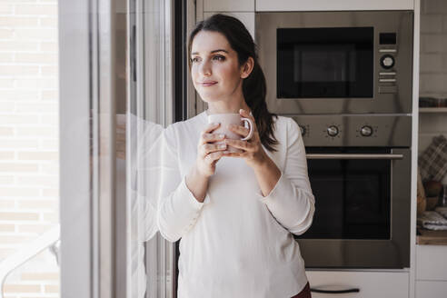 Thoughtful woman with coffee cup standing near window - EBBF08154