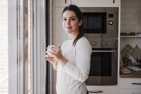 Lächelnde Frau mit Kaffeetasse am Fenster stehend - EBBF08155