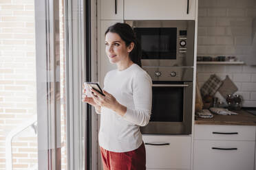 Lächelnde Frau mit Smartphone und Kaffeetasse, die durch ein Fenster in der Küche schaut - EBBF08156