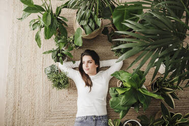 Nachdenkliche Frau entspannt sich bei Pflanzen auf dem Teppich zu Hause - EBBF08158