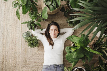 Lächelnde Frau mit Händen hinter dem Kopf, die sich auf einem Teppich inmitten von Pflanzen entspannt - EBBF08159