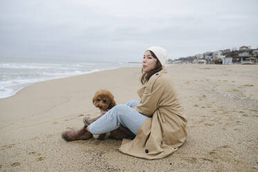 Junge Frau mit Maltipoo-Hund sitzt am Strand im Sand - ALKF00171