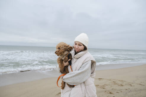 Lächelnde Frau in warmer Kleidung mit Hund am Strand - ALKF00178