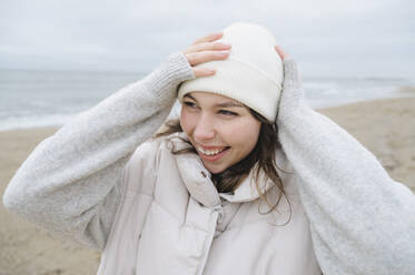 Happy woman adjusting knit hat at beach - ALKF00182