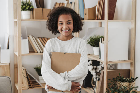 Smiling girl holding spiral notebook at home - EBBF08164