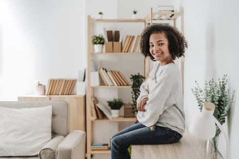 Smiling girl with curly hair sitting on desk at home - EBBF08166