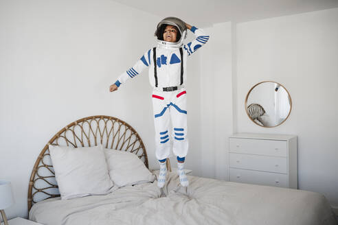 Happy girl wearing space suit jumping on bed at home - EBBF08170