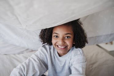 Happy girl sitting on bed under blanket at home - EBBF08172