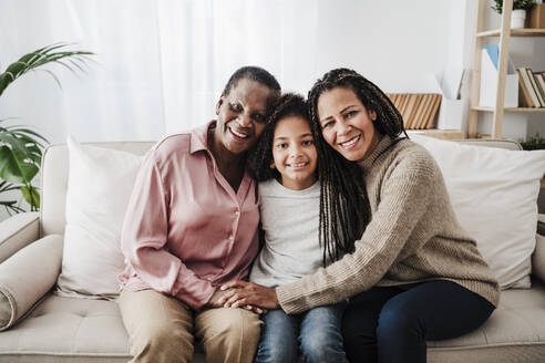 Smiling woman sitting with mother and daughter on sofa at home - EBBF08196