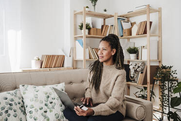 Thoughtful woman with laptop sitting on sofa at home - EBBF08218