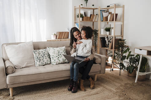 Smiling girl and mother sitting with laptop on sofa at home - EBBF08220