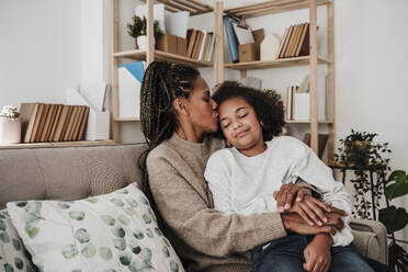 Mother kissing daughter sitting on sofa at home - EBBF08222