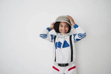 Smiling girl adjusting space helmet in front of white wall - EBBF08238