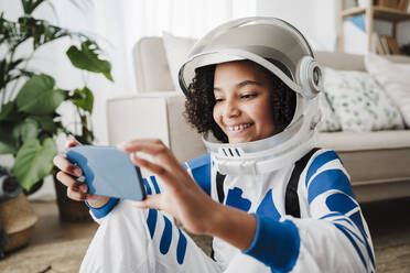 Mädchen im Raumanzug mit Smartphone zu Hause sitzend - EBBF08249