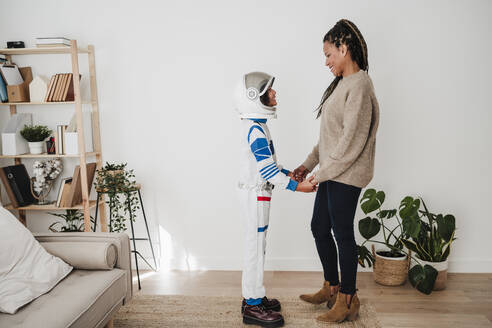 Smiling woman standing with daughter wearing space suit near wall at home - EBBF08261