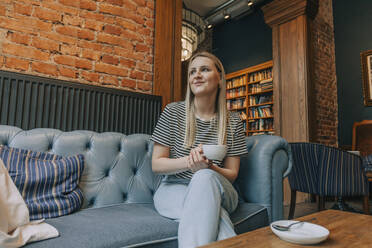 Lächelnde Frau sitzt mit einer Kaffeetasse auf einer Couch in einem Cafe - VSNF00613