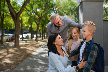 Glückliche Großeltern, die ihre Enkelkinder von der Schule nach Hause bringen und vor einer Schule im Freien auf der Straße warten. - HPIF08323
