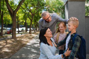 Happy grandparents taking grandchildren home from school, waiting in front of a school outdoors in street. - HPIF08323