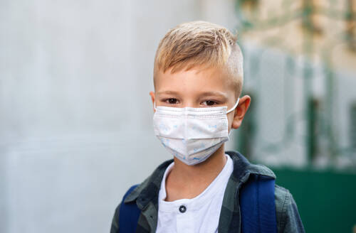 A happy little schoolboy with face mask looking at camera in front of school. - HPIF08345