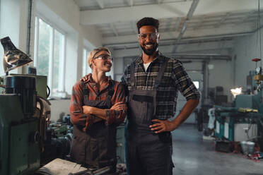 Ein Porträt von jungen, gemischtrassigen Arbeitskollegen, die in einer Metallwerkstatt arbeiten, lächeln und in die Kamera schauen. - HPIF08354