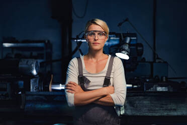 A portrait of mid adult woman standing with arms crossed indoors in metal workshop, looking at camera. - HPIF08371