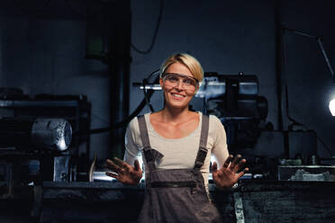 Portrait of a happy young industrial woman with dirty hands looking at camera indoors in metal workshop. - HPIF08379