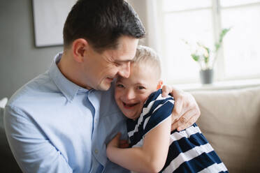 A father with happy down syndrome son indoors at home, hugging and laughing. - HPIF08402