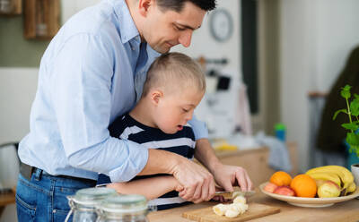 A father with happy down syndrome son indoors in kitchen, chopping fruit. - HPIF08406