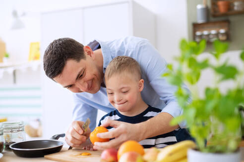 A father with happy down syndrome son indoors in kitchen, preparing food. - HPIF08408