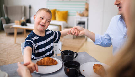 Ein Porträt eines glücklichen Jungen mit Down-Syndrom, der mit seinen Eltern am Tisch sitzt und frühstückt. - HPIF08426