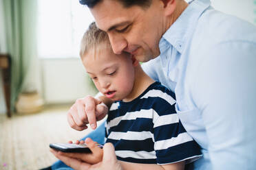 A father with happy down syndrome son indoors at home, using smartphone. - HPIF08438