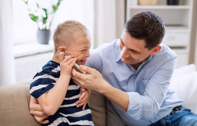 A father with happy down syndrome son indoors at home, using smartphone. - HPIF08442