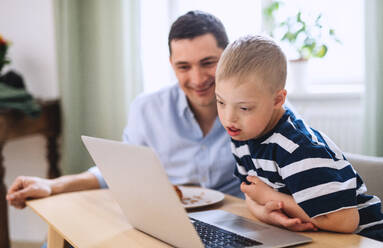 Ein Vater mit einem glücklichen Sohn mit Down-Syndrom, der zu Hause einen Laptop benutzt. - HPIF08447