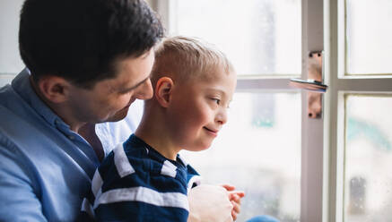 Älterer Vater mit glücklichem Sohn mit Down-Syndrom zu Hause, der durch das Fenster schaut. - HPIF08450
