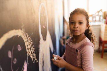 Ein kleines Mädchen malt mit Kreide auf einer Tafel im Spielzimmer und schaut in die Kamera. - HPIF08517