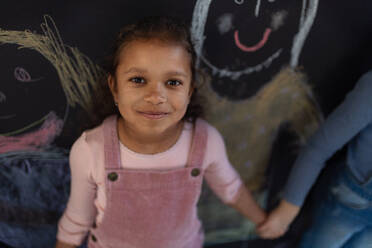 Kleines Mädchen posiert und lächelnd vor der Tafel Wand Zeichnungen drinnen im Spielzimmer. - HPIF08523