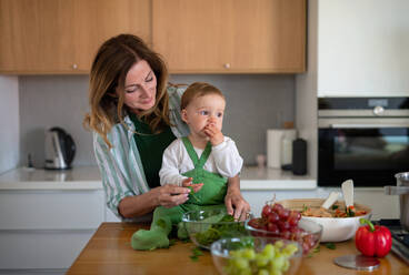 Portrait of happy mature woman with small baby granddaughter indoors at home cooking. - HPIF08539