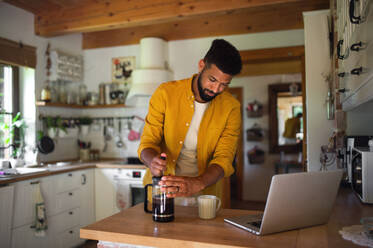 Ein junger Mann mit Laptop und Kaffee arbeiten drinnen, Home-Office-Konzept. - HPIF08601