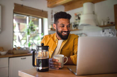 Ein junger Mann mit Laptop und Kaffee arbeiten drinnen, Home-Office-Konzept. - HPIF08602