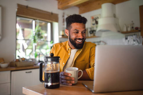 A young man with laptop and coffee working indoors, home office concept. - HPIF08602