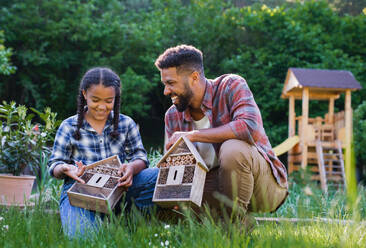 Ein glücklicher junger Mann mit einer kleinen Schwester, die lachend Käferhotels im Garten hält. - HPIF08618