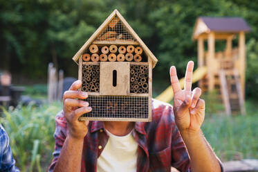 Frontansicht eines glücklichen jungen Mannes mit einem Insektenhotel, der seine Zeit im Freien im Garten verbringt und mit den Fingern das V-Zeichen zeigt. - HPIF08620