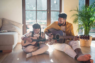 A front view of happy father with small daughter indoors at home, playing guitar. - HPIF08651