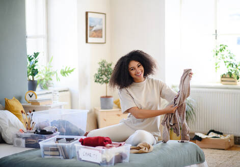 Happy young woman sorting clothing indoors at home, charity donation concept. - HPIF08684