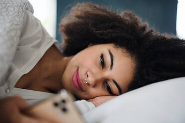 Porträt einer glücklichen jungen Frau auf dem Bett, die ein Smartphone benutzt. - HPIF08722