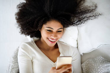 Top view portrait of happy young woman indoors on bed, using smartphone. - HPIF08723