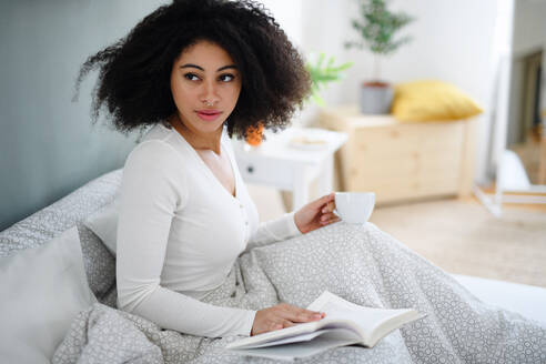 Close-up portrait of happy young woman with book and coffee indoors, relaxing in bed. - HPIF08726